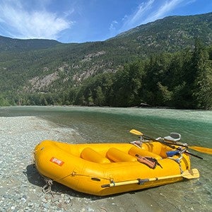 A yellow raft sitting on the bank of a river backed by a wooded mountain range.