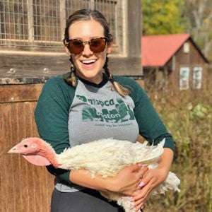 A woman wearing sunglasses smiles at the camera while holding a live adult turkey