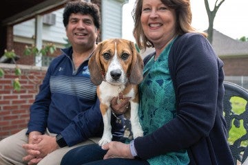 Teddy, a medium-sized beagle, stares into the camera while sitting with his adopters, Greta Guest and Dave Rubello